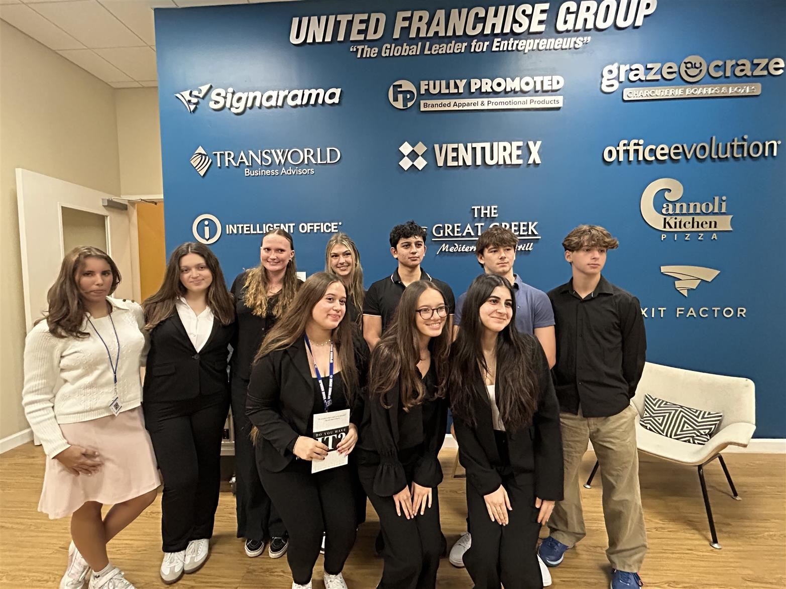  A group of business choice students posing in front of a United Franchise Group wall of businesses